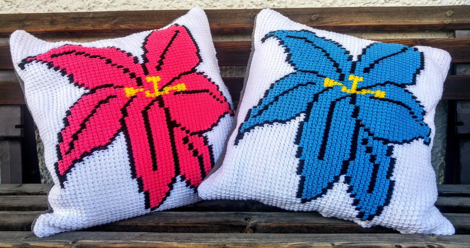 Zwei handgehäkelte Kissenbezüge mit großen Blumenmotiven in Rosa und Blau auf Holzbank