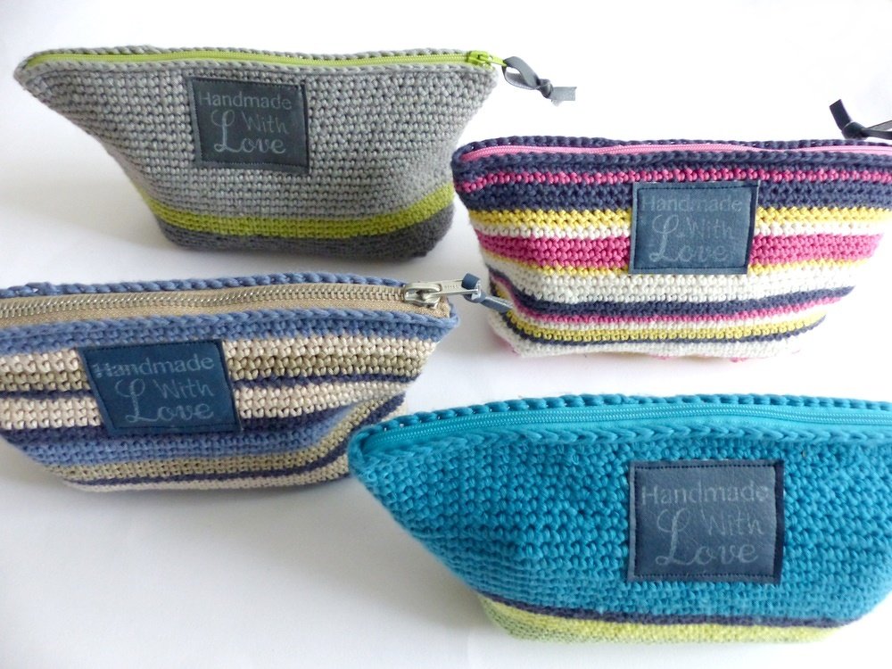 Vier gehäkelte Reißverschlusstaschen in verschiedenen Farben und Streifen mit ‚Handmade With Love‘-Etiketten