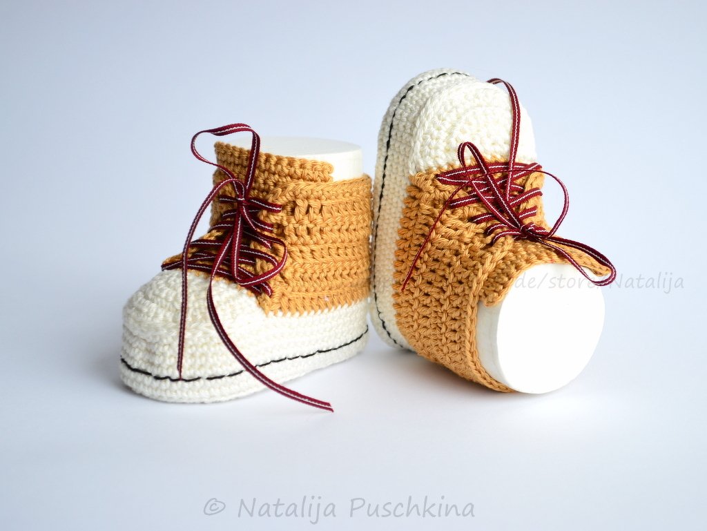 Ein Paar gehäkelter Babyschuhe im High‑Top‑Sneaker‑Look aus Senfgelb und Creme mit dunkelroten Schnürsenkeln.