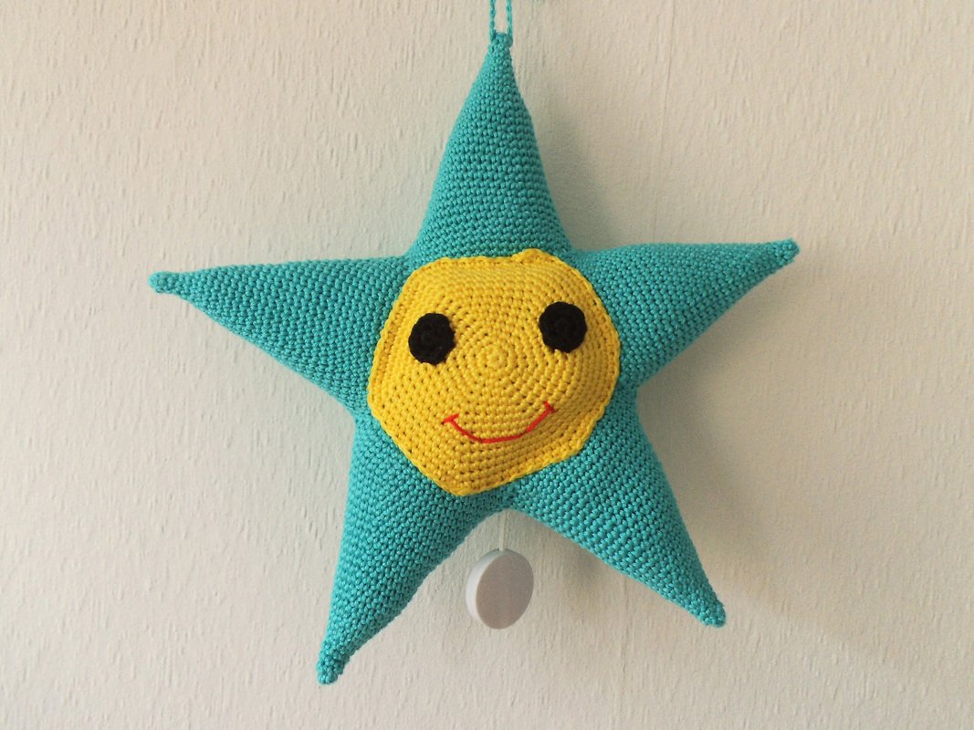 Türkis-gelber gehäkelter Stern mit gelbem Smiley und weißem Zugring vor heller Wand