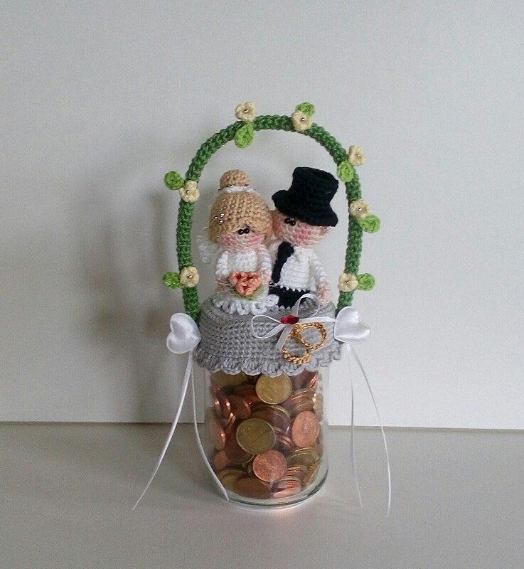 Gehäkeltes Brautpaar mit Zylinder und Schleier auf einem mit Münzen gefüllten Glas, dekoriert mit einem grünen Blumenbogen.