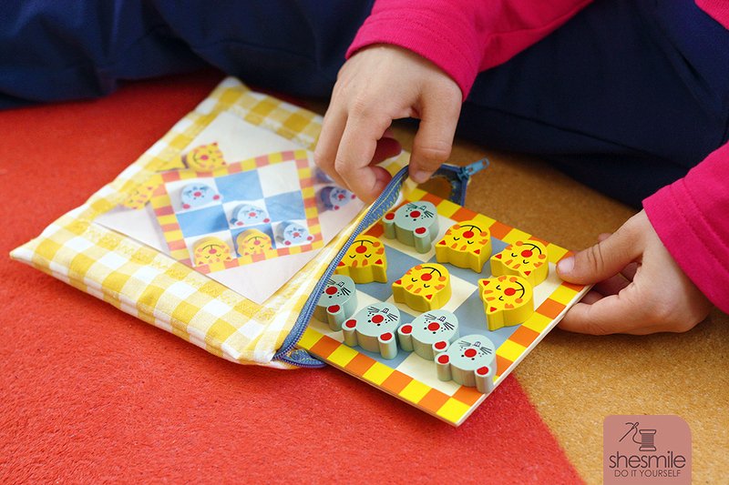 Flaches kariertes Utensilo mit Reißverschluss und Sichtfenster, darin ein Spielbrett mit Holzfiguren; Kinderhände greifen hinein.