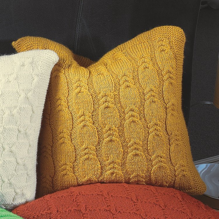 Senfgelbes Strickkissen mit Zopfmuster auf dunklem Sofa, daneben weiße und rote Kissen