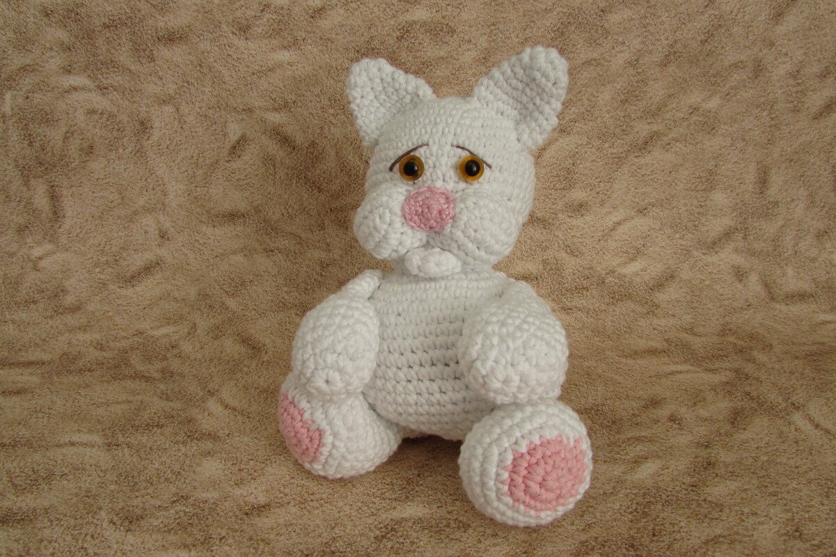 Weißes gehäkeltes Amigurumi-Kätzchen mit rosa Nase und rosa Pfotenpads sitzt auf beigem Stoff