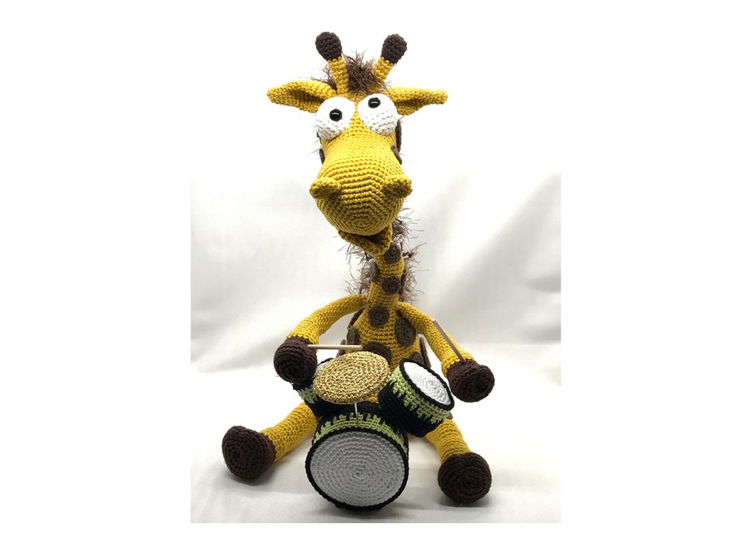 Gelbe gehäkelte Amigurumi-Giraffe mit braunen Flecken, die auf einem weißen Hintergrund sitzt und drei kleine Trommeln hält.