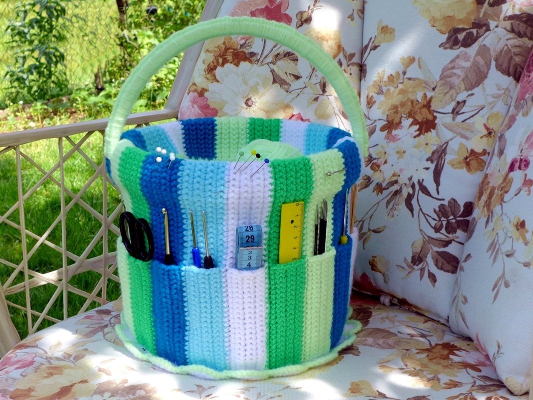 Blue, green and white striped crocheted basket with exterior pockets holding scissors, crochet hooks and measuring tape