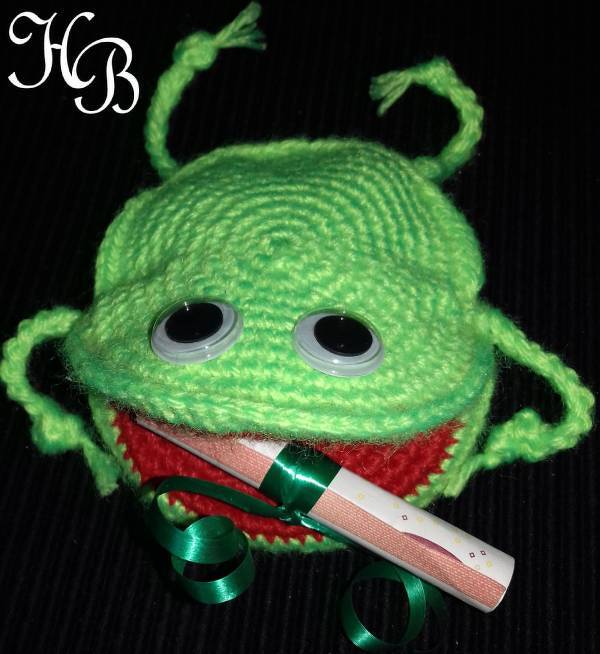 Grünes gehäkeltes Amigurumi‑Monster mit Wackelaugen, offenem roten Maul und gerollter Papierrolle