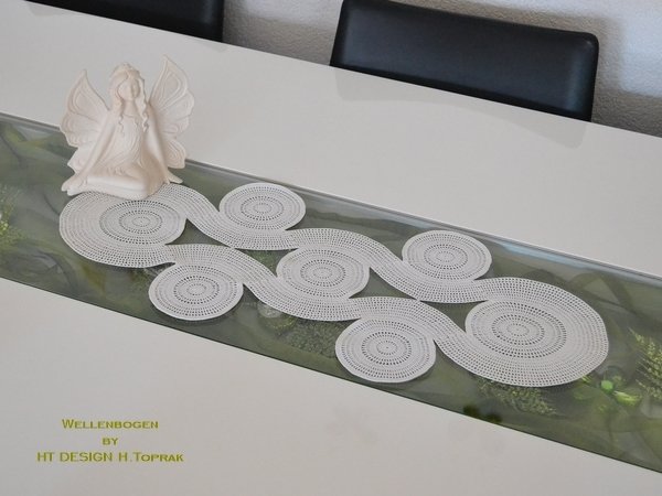 White crocheted table runner with circular wave motifs on a white table and glass center panel, with a small angel figurine.