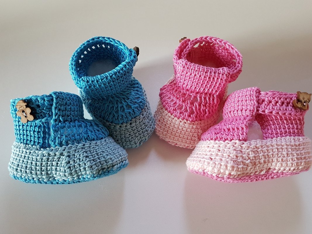 Zwei Paar gehäkelte Babyschuhe in Blau und Rosa mit kleinen Holzknöpfen auf hellem Untergrund.