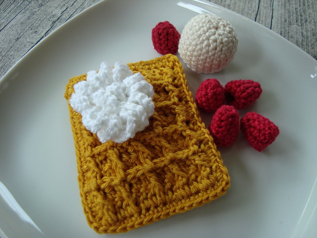 Gehäkelte goldgelbe Waffel mit weißer Sahne, rote gehäkelte Himbeeren und eine weiße gehäkelte Kugel auf einem weißen Teller.