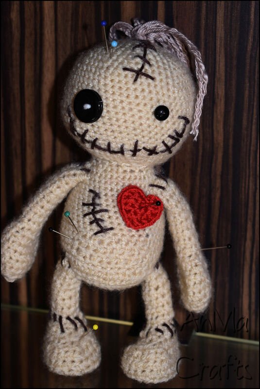 Beige gehäkelte Amigurumi-Puppe mit Knopfaugen, gesticktem Mund, rotem Herz und bunten Stecknadeln