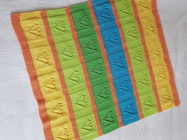 Nahaufnahme einer gestrickten Babydecke mit Streifen in Gelb, Grün, Blau und Orange; erhabenes Blättermuster in den farbigen Feldern.