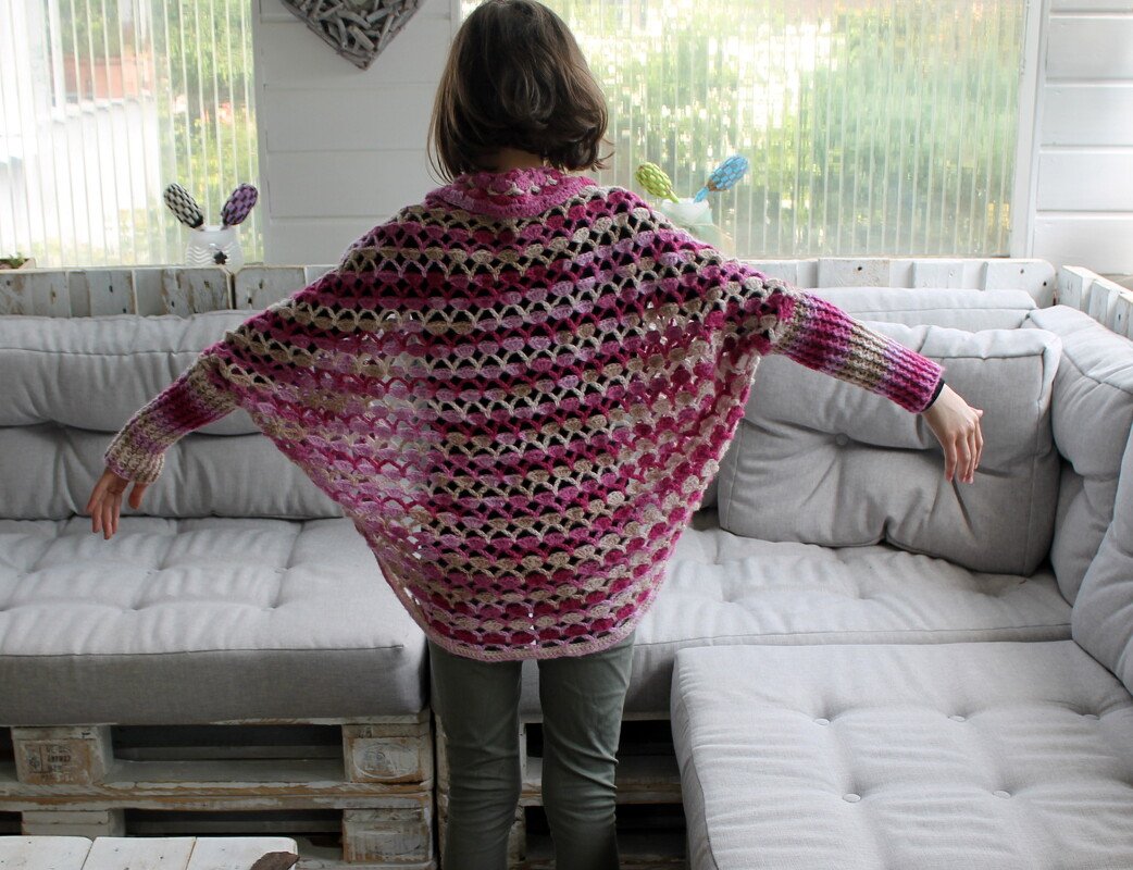 Rückenansicht einer Person trägt gehäkelten Seelenwärmer in Rosa- und Naturtönen mit offenem Muschelmuster und langen Bündchen vor hellem Sofa