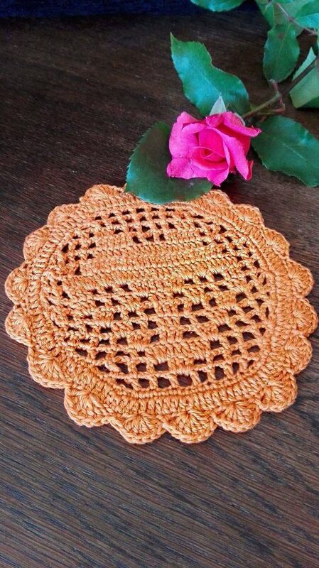 Rundes rostorangefarbenes Häkeldeckchen mit Ajourmuster und Muschelrand, darauf eine pinke Rose auf dunklem Holztisch