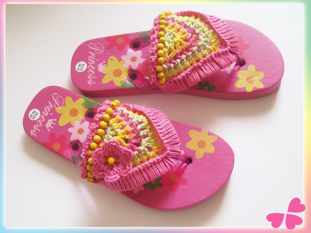Zwei pinke Kinderflip-Flops mit bunten gehäkelten Riemenverzierungen, gelben Perlen und kleinen Blumen auf weißem Hintergrund.