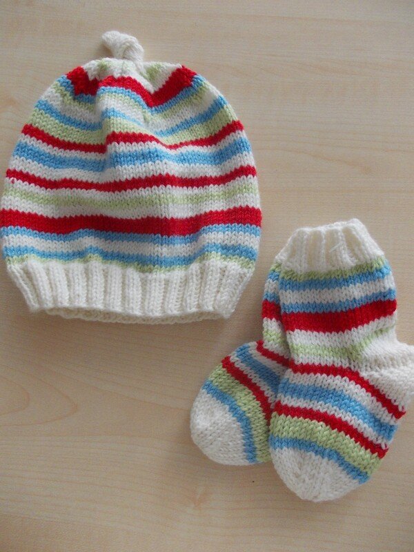Gestricktes Babyset mit Ringelstreifen: Mütze mit kleinem Knoten und Söckchen in Rot, Hellblau, Hellgrün und Creme auf hellem Holzuntergrund.