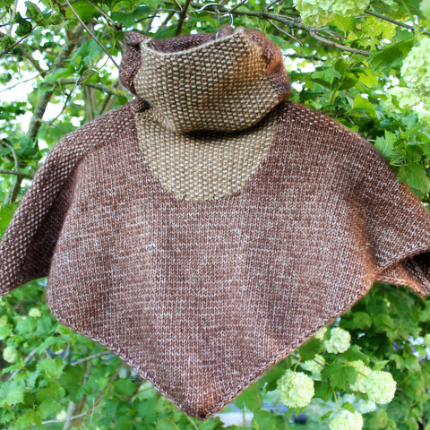 Gestrickter Kinderponcho in Braun und Beige mit strukturiertem Rollkragen vor grünen Blättern.