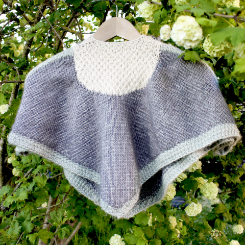 Gestrickter Kinderponcho grau-mint mit strukturierter Passe an Bügel vor blühendem Strauch
