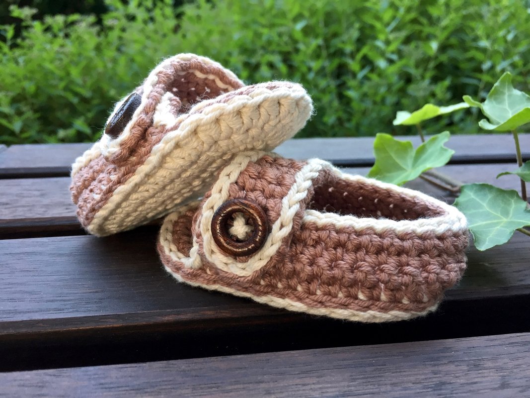 Zwei gehäkelte Babyschuhe in Beige und Braun mit Holzknöpfen auf dunklem Holz