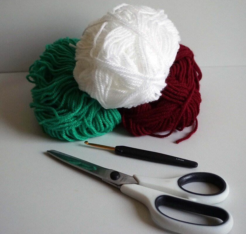 Green, white and red yarn with a crochet hook and scissors on a white surface.