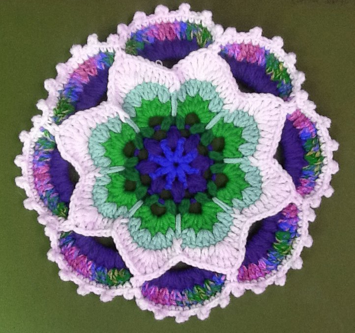 Rundes gehäkeltes Mandala in Blütenform mit Weiß, Violett, Grün, Blau und Rosa auf grünem Hintergrund.