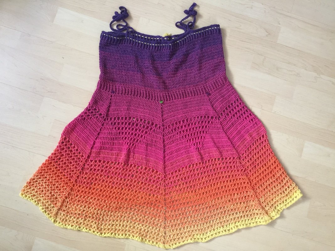 Gehäkeltes Damenkleid mit Farbverlauf (Lila–Pink–Orange–Gelb) auf hellem Holzboden liegend.