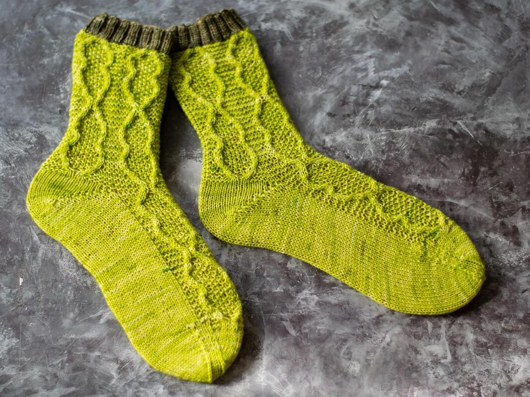 Zwei handgestrickte lindgrüne Socken mit Zopfmuster und dunkelolivgrünen Rippenbündchen auf grauem, strukturiertem Untergrund.