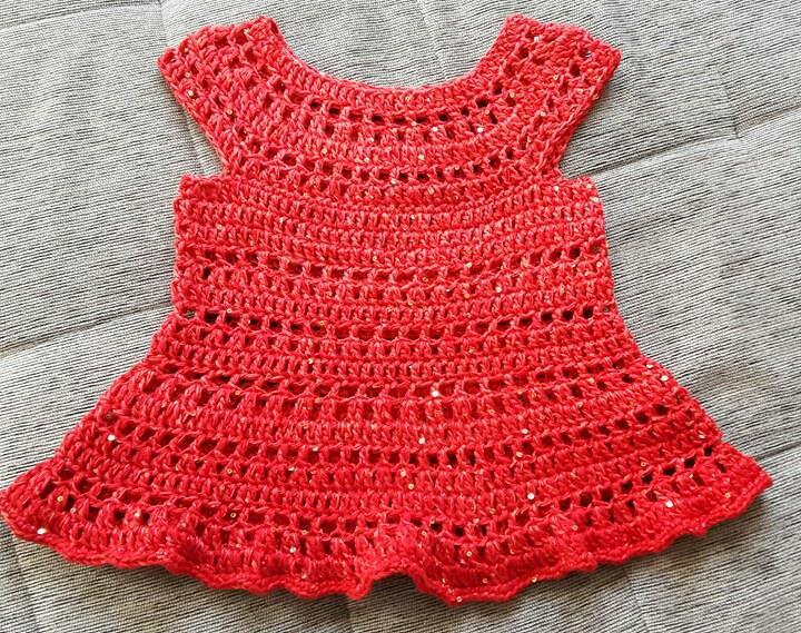 Rotes gehäkeltes Babykleid mit offenem Muster und dezenten Glitzereffekten auf hellem Hintergrund.