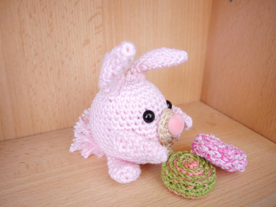 Rosa gehäkeltes Amigurumi-Häschen mit schwarzen Augen und zwei gehäkelten Dekorationen auf Holzuntergrund