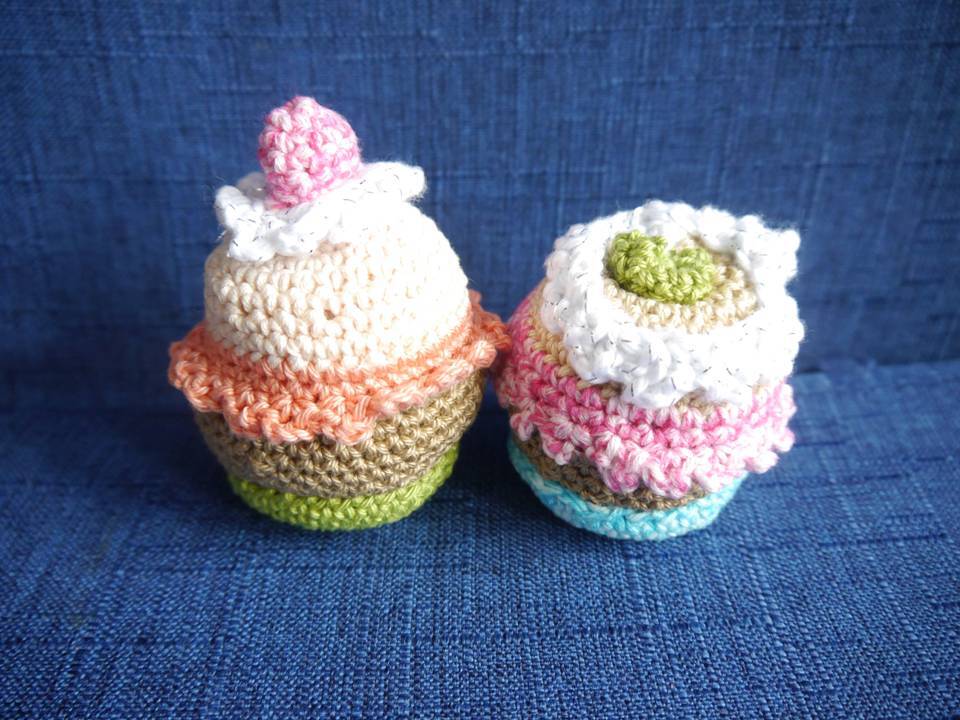 Zwei gehäkelte Cupcakes in Pastellfarben auf blauem Stoff, einer mit pinker Deko, einer mit grünem Detail.