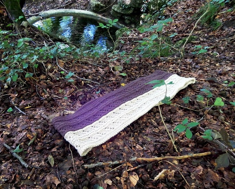 Purple and cream cable and lace knitted loop scarf laid flat on leaf-strewn forest floor