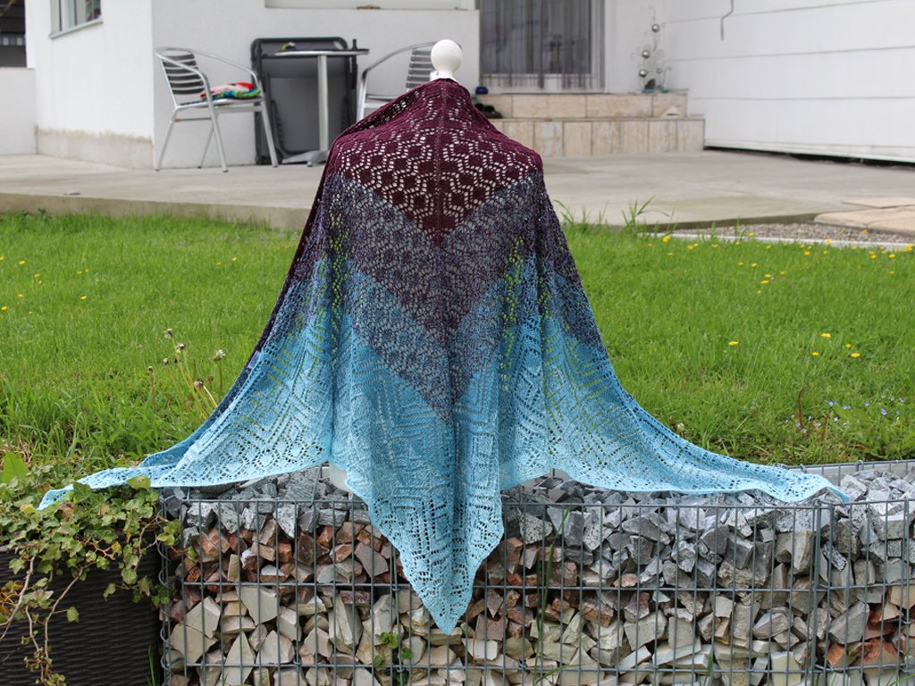 Back view of a knitted ombre triangle shawl with lace stitch patterns, transitioning from burgundy to turquoise, draped on a mannequin outdoors.