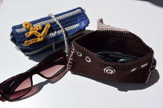 Zwei gehäkelte Brillenetuis: blau-weiß gestreift mit Anker und braun mit Metallösen; eine Sonnenbrille liegt daneben.