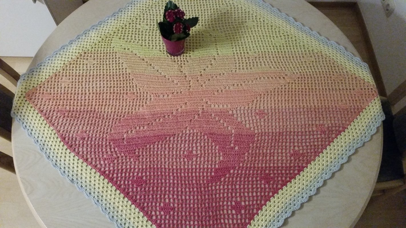 Quadratisches handgehäkeltes Deckchen mit Farbverlauf von Gelb zu Pink und hellem Wellenrand auf einem Tisch, dekoriert mit einem kleinen Blumentopf.