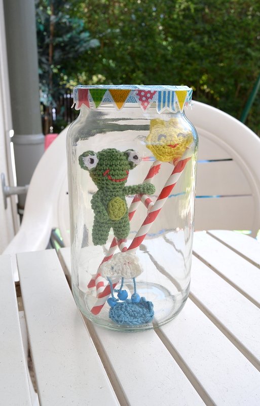 Gehäkelter grüner Frosch, gelbe Sonne und Wolke mit Tropfen in einem Glas mit rot-weiß gestreiften Strohhalmen.