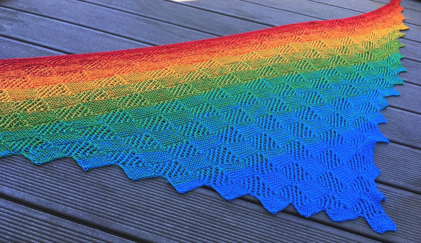 Gestricktes Dreieckstuch mit Regenbogen-Farbverlauf, feinem Lochmuster und gezacktem Rand auf dunklem Holz.