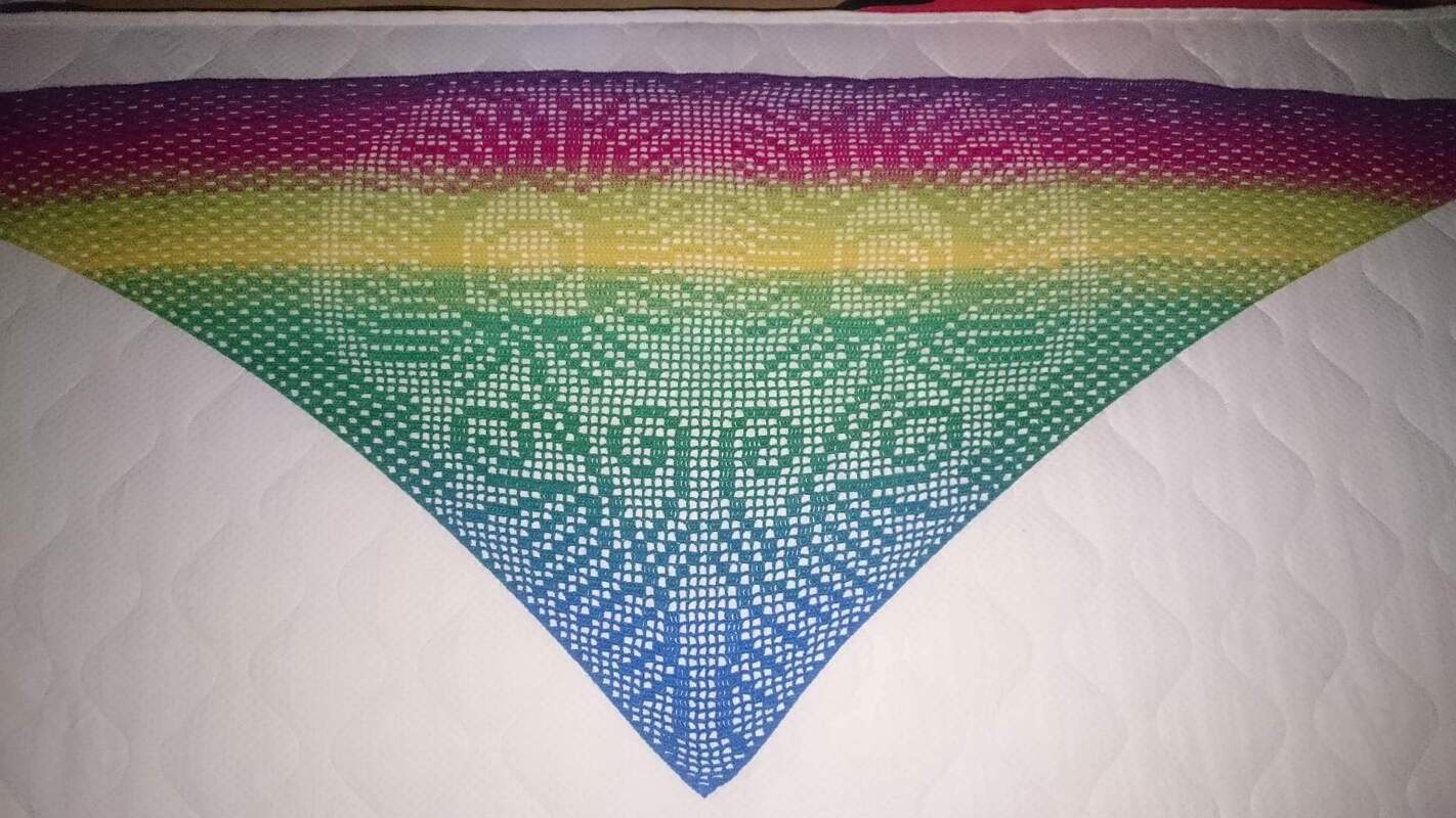 Gehäkeltes dreieckiges Tuch mit Farbverlauf von Violett über Gelb-Grün zu Blau und filigranem Lochmuster auf hellem Untergrund.