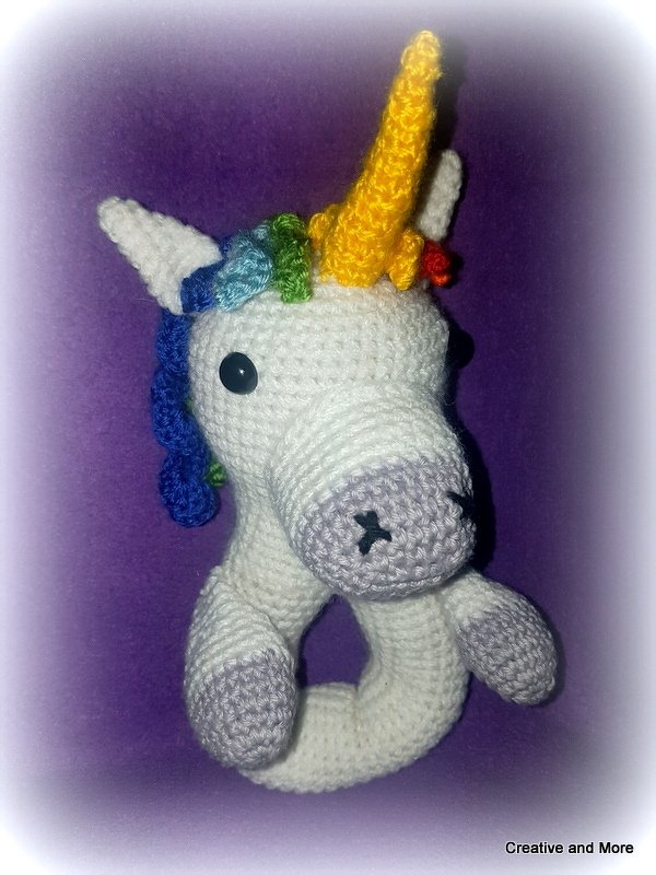 Gehäkelte weiße Einhorn-Rassel mit gelber Hornspitze und regenbogenfarbener Mähne vor lila Hintergrund.