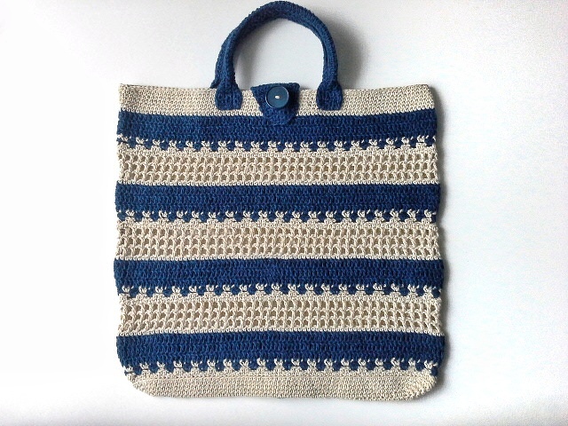 Handgehäkelter Shopper mit horizontalen Streifen in Marineblau und Beige, blauen Tragegriffen und Knopfverschluss auf weißem Hintergrund.
