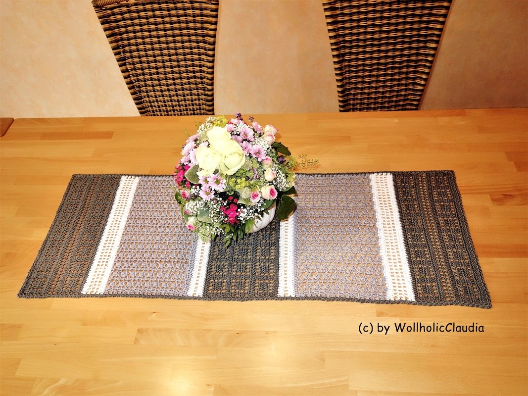 Gehäkelter Tischläufer mit Streifen in Grau, Anthrazit und Weiß auf hellem Holztisch, mittig dekoriert mit einem Blumenstrauß.