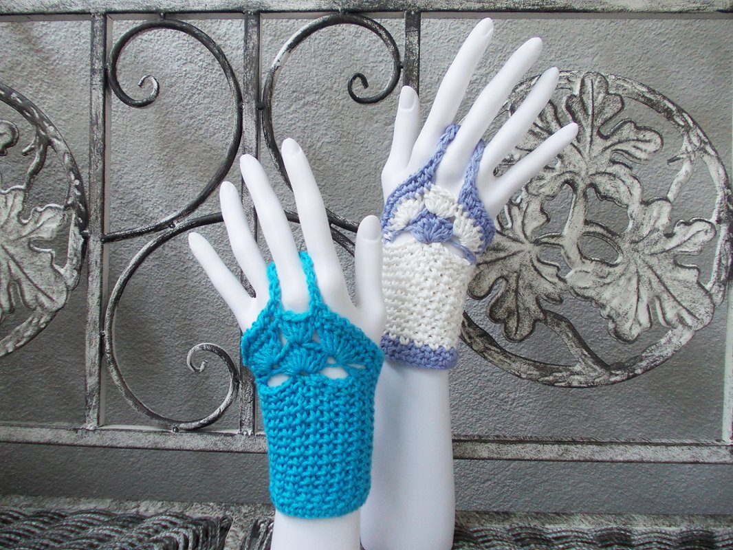 Zwei gehäkelte fingerlose Handschuhe in Türkis und Weiß-Lila auf Mannequin-Händen vor ornamentalem Metallgitter