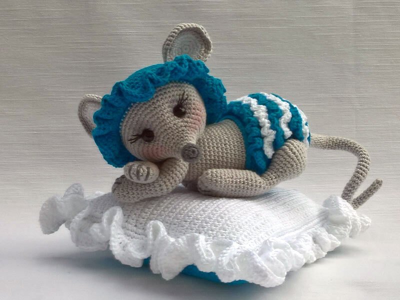 Beige crocheted amigurumi mouse with teal ruffled bonnet and diaper lying on a white ruffled pillow