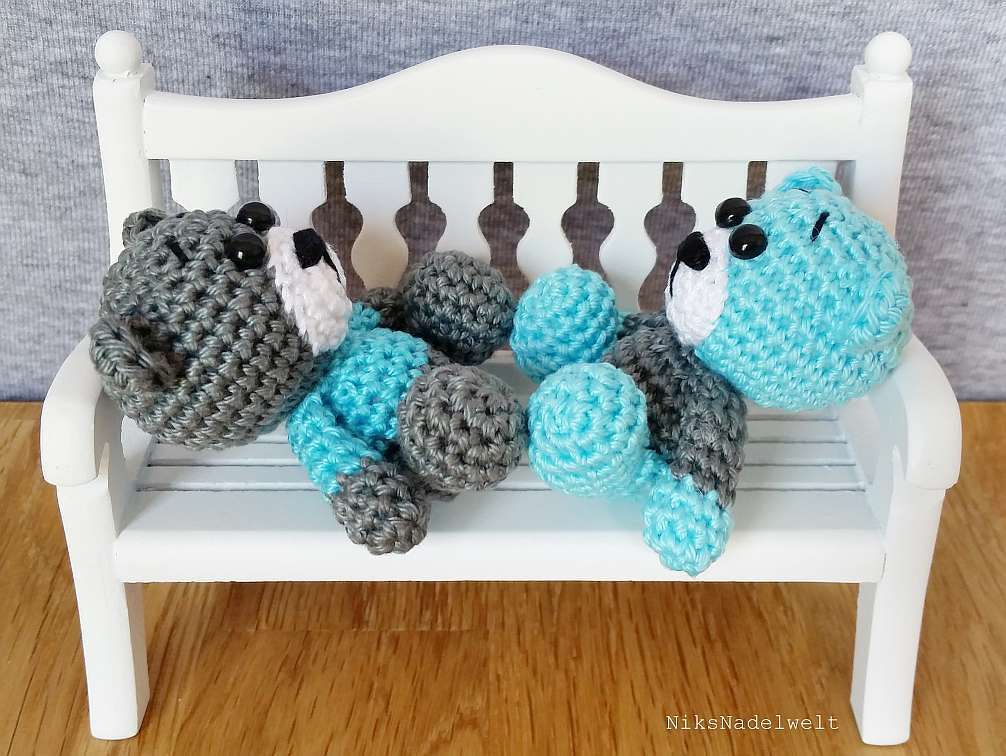 Handgehäkelte Amigurumi-Bären in Grau und Türkis auf einer kleinen weißen Bank.