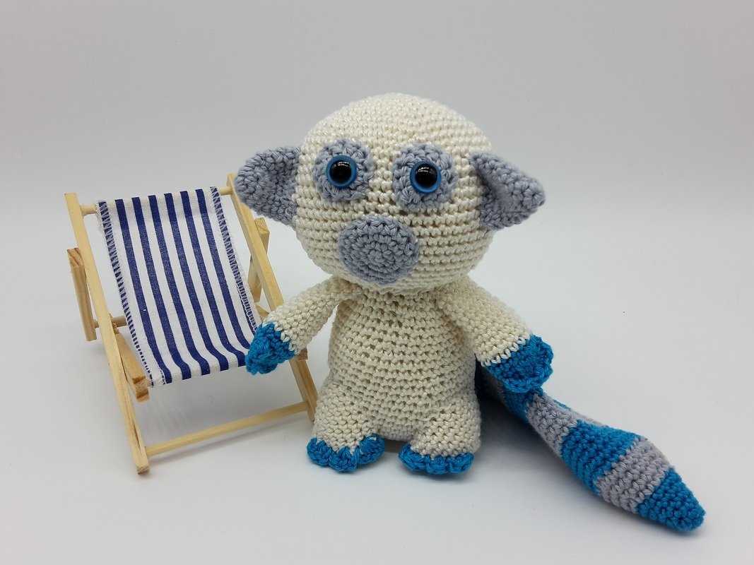 Gehäkeltes Lemur-Amigurumi in Creme, Grau und Blau mit geringeltem Schwanz neben kleinem blau-weiß gestreiftem Liegestuhl