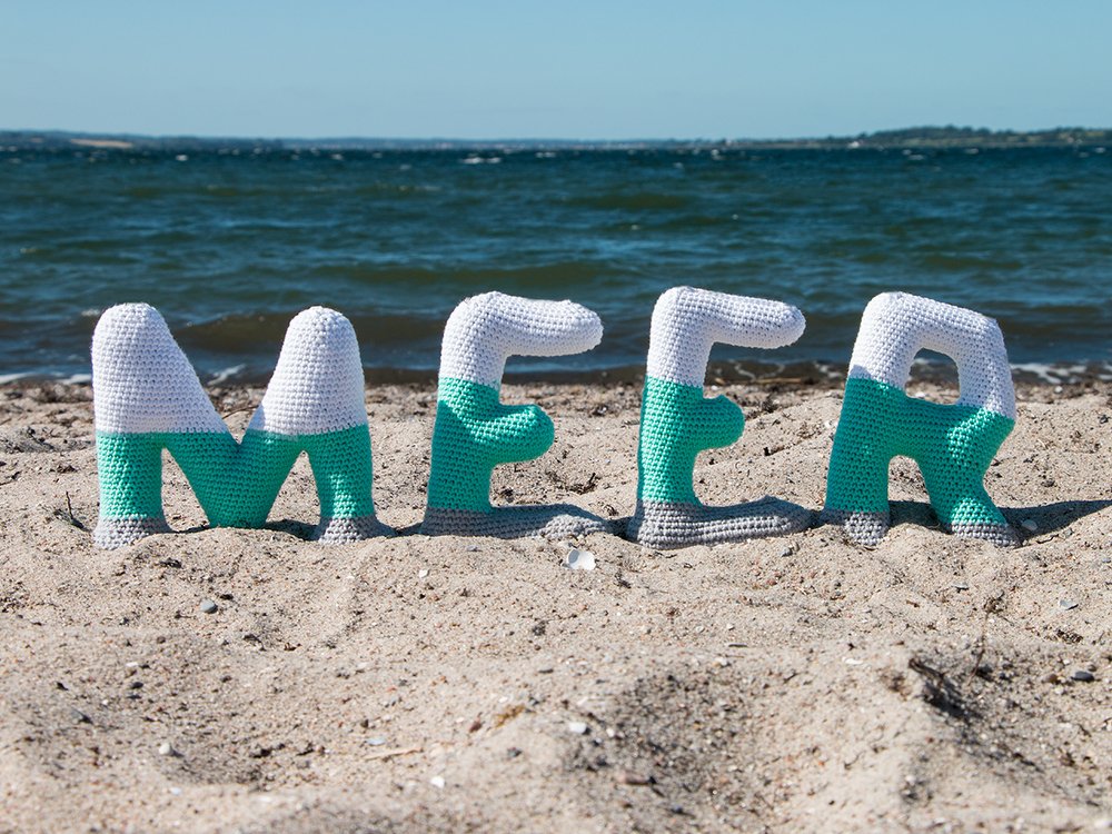Gehäkelte Buchstaben 'MEER' in Weiß, Türkis und Grau auf Sandstrand vor dem Meer