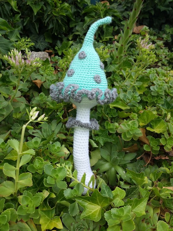 Gehäkelter Pilz mit mintgrüner Kappe, grauen Punkten und weißem Stiel zwischen Pflanzen