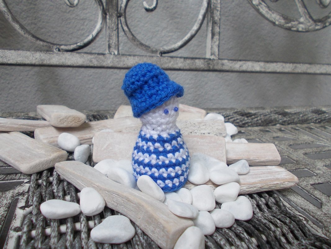 Gehäkelte Amigurumi-Figur mit blauem Hut und blau-weißem Streifenmuster, umgeben von Holzstücken und weißen Kieseln.