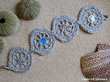 Gehäkeltes Armband aus silbergrauem Garn mit runden Motiven auf Sand, umgeben von Seeigelgehäusen.