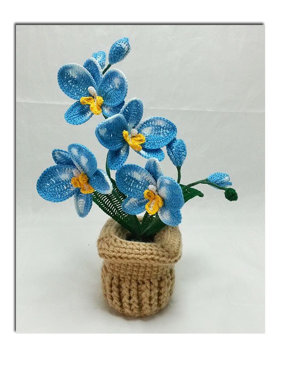 Gehäkelte blaue Orchidee mit gelben Zentren und grünen Blättern in beigem Häkeltopf vor hellem Hintergrund.