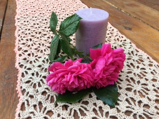 Cremefarbener gehäkelter Tischläufer mit Blütenmuster, pinke Rosen und pastelllila Kerze auf Holztisch